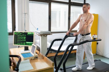Orta yaşlı bir adam, klinik bir ortamda kalp izleme için EKG elektrotlarına bağlı bir koşu bandında tıbbi stres testinden geçiyor.. 