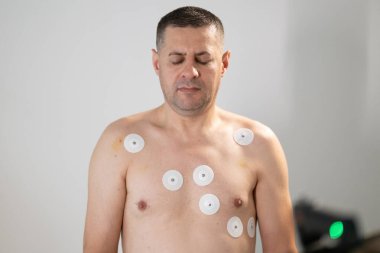 Orta yaşlı bir adam, klinik bir ortamda kalp izleme için EKG elektrotlarına bağlı bir koşu bandında tıbbi stres testinden geçiyor.. 
