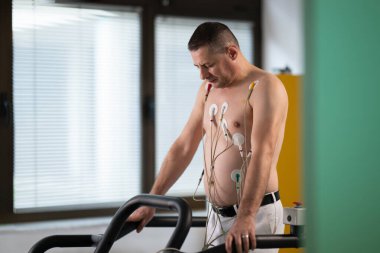 Orta yaşlı bir adam, klinik bir ortamda kalp izleme için EKG elektrotlarına bağlı bir koşu bandında tıbbi stres testinden geçiyor.. 