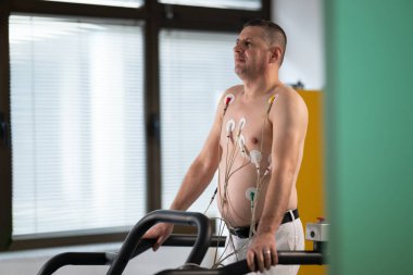 Orta yaşlı bir adam, klinik bir ortamda kalp izleme için EKG elektrotlarına bağlı bir koşu bandında tıbbi stres testinden geçiyor.. 