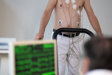 Orta yaşlı bir adam, klinik bir ortamda kalp izleme için EKG elektrotlarına bağlı bir koşu bandında tıbbi stres testinden geçiyor.. 