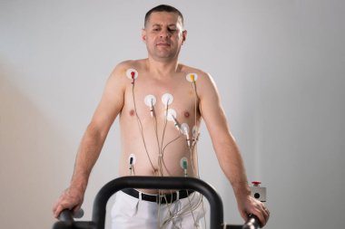 Orta yaşlı bir adam, klinik bir ortamda kalp izleme için EKG elektrotlarına bağlı bir koşu bandında tıbbi stres testinden geçiyor.. 
