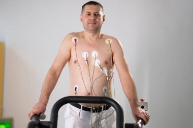 Orta yaşlı bir adam, klinik bir ortamda kalp izleme için EKG elektrotlarına bağlı bir koşu bandında tıbbi stres testinden geçiyor.. 