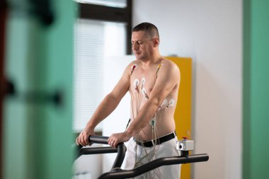 Orta yaşlı bir adam, klinik bir ortamda kalp izleme için EKG elektrotlarına bağlı bir koşu bandında tıbbi stres testinden geçiyor.. 