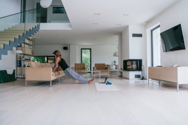 Evde antrenman. Sportif adam antrenman yaparken dizüstü bilgisayarda özel ders izliyor oturma odasında egzersiz yapıyor, boş yer var.