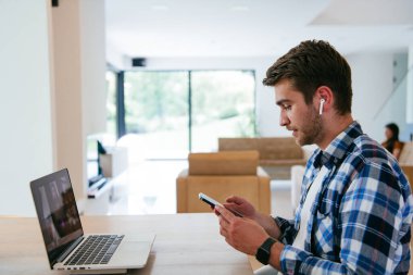 Modern bir oturma odasındaki masada oturan bir serbest çalışan, kulaklıklarıyla iş görüşmesi için bir laptop kullanan, arkadaşlarıyla sohbet eden ve eğlenen biri..