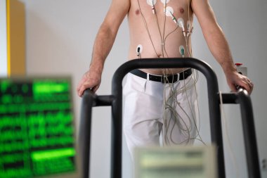 Orta yaşlı bir adam, klinik bir ortamda kalp izleme için EKG elektrotlarına bağlı bir koşu bandında tıbbi stres testinden geçiyor.. 