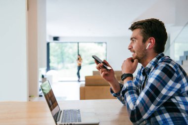 Modern bir oturma odasındaki masada oturan bir serbest çalışan, kulaklıklarıyla iş görüşmesi için bir laptop kullanan, arkadaşlarıyla sohbet eden ve eğlenen biri..