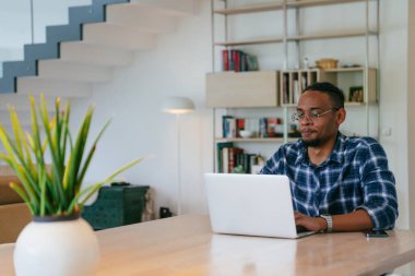 Modern bir oturma odasındaki masada oturan gözlüklü Afrikalı Amerikalı etkileyiciler iş görüşmesi, arkadaşlarla sohbet ve eğlence için dizüstü bilgisayar kullanıyorlar.