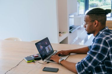 Modern bir oturma odasındaki masada oturan gözlüklü Afrikalı Amerikalı etkileyiciler iş görüşmesi, arkadaşlarla sohbet ve eğlence için dizüstü bilgisayar kullanıyorlar.