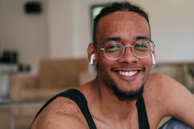 Afrika kökenli Amerikalı bir adamın gözlüklü ve kulaklıklı yüzü ciddi sivilcelerle kaplı oturma odasında yerde otururken fotoğrafı.