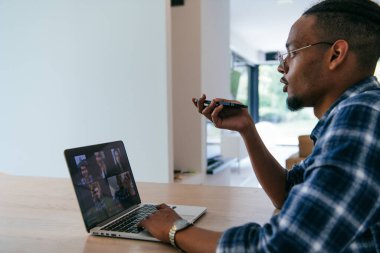 Modern bir oturma odasındaki masada oturan gözlüklü Afrikalı Amerikalı iş adamı bilgisayarını ve akıllı telefonunu iş videosu sohbeti, arkadaşlarla sohbet ve eğlence için kullanıyor.