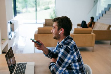 Modern bir oturma odasındaki masada oturan bir serbest çalışan, kulaklıklarıyla iş görüşmesi için bir laptop kullanan, arkadaşlarıyla sohbet eden ve eğlenen biri..