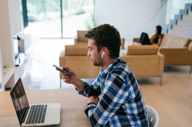 Modern bir oturma odasındaki masada oturan bir serbest çalışan, kulaklıklarıyla iş görüşmesi için bir laptop kullanan, arkadaşlarıyla sohbet eden ve eğlenen biri..