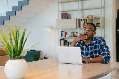 Modern bir oturma odasındaki masada oturan gözlüklü Afrikalı Amerikalı etkileyiciler iş görüşmesi, arkadaşlarla sohbet ve eğlence için dizüstü bilgisayar kullanıyorlar.