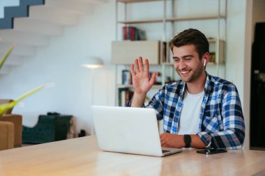 Modern bir oturma odasındaki masada oturan bir iş adamı, kulaklıklarıyla iş videosu sohbeti, arkadaşlarla sohbet ve eğlence için dizüstü bilgisayar kullanıyor.