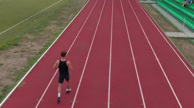 İşaretli bir yarış pisti boyunca koşan, pistin sınırlarını gösteren, hız, odaklanma ve hareket halindeki hassasiyeti vurgulayan tek kişilik bir erkek atletin havadan üst görüntüsü. Sinematik İHA yavaş çekim