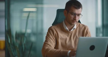 Akıllı görünümlü odaklanmış iş adamı bir dizüstü bilgisayarda çalışıyor ve düşünüyor. Milenyum dönemi aile bütçesini yönetmek, finansal evrak işlerini halletmek ve modern bir açık uzay ofisindeki dizüstü bilgisayardaki harcamaları takip etmek.