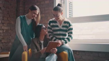 Kadın iş adamları pencere kenarında iş birliği yapıyor, fikirleri tartışıyor ve tablet üzerinde beyin fırtınası yapıyorlar. Modern bir ofiste işbirliği, yaratıcılık ve takım çalışması kavramı