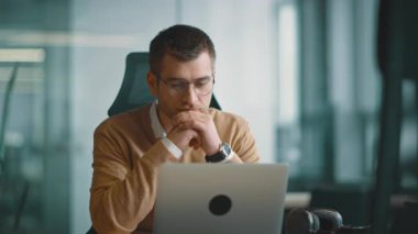 Akıllı görünümlü odaklanmış iş adamı bir dizüstü bilgisayarda çalışıyor ve düşünüyor. Milenyum dönemi aile bütçesini yönetmek, finansal evrak işlerini halletmek ve modern bir açık uzay ofisindeki dizüstü bilgisayardaki harcamaları takip etmek.