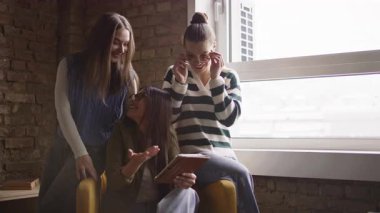 Kadın iş adamları pencere kenarında iş birliği yapıyor, fikirleri tartışıyor ve tablet üzerinde beyin fırtınası yapıyorlar. Modern bir ofiste işbirliği, yaratıcılık ve takım çalışması kavramı