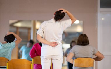 Bir fizyoterapist bir grup hastaya boyun esnetme ve tam vücut rehabilitasyon egzersizlerinde rehberlik eder, hareket kabiliyetlerini, esnekliklerini ve genel iyileşme süreçlerini geliştirmelerine yardımcı olur. 