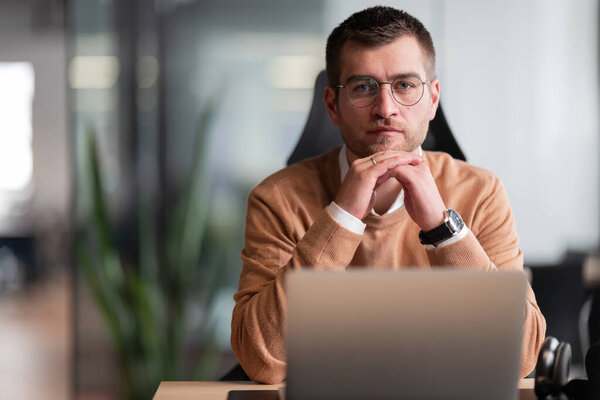 Smart looking focused businessman working on a laptop and thinking. Millennial managing family budget, handling financial paperwork, and tracking expenditures on a laptop at a modern open space office