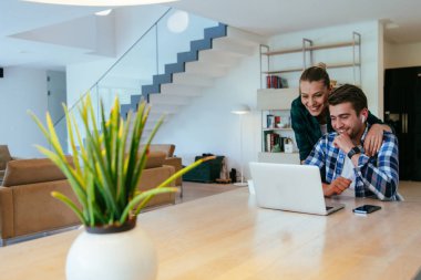 Genç evli bir çift, modern evlerinin oturma odasında otururken bir dizüstü bilgisayarla ebeveynleri, aileleri ve arkadaşlarıyla konuşuyor.