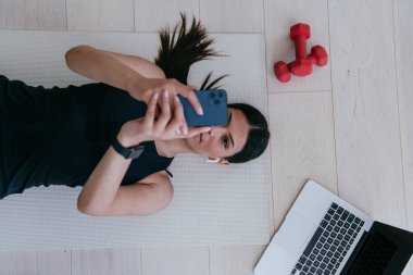 Sporcu kadın, modern evin zemininde dinlenirken akıllı telefon kullanıyor.