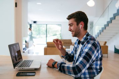 Modern bir oturma odasındaki masada oturan bir serbest çalışan, kulaklıklarıyla iş görüşmesi için bir laptop kullanan, arkadaşlarıyla sohbet eden ve eğlenen biri..