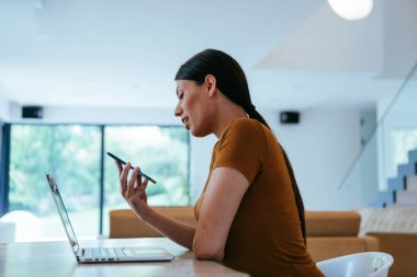 Oturma odasında dizüstü bilgisayar ve akıllı telefon kullanarak oturan bir kadın iş arkadaşları ve akrabalarıyla yaptığı görüntülü konuşmaya bakıyor. İş görüşmesi soruları yanıtlıyor