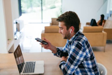 Modern bir oturma odasındaki masada oturan bir serbest çalışan, kulaklıklarıyla iş görüşmesi için bir laptop kullanan, arkadaşlarıyla sohbet eden ve eğlenen biri..