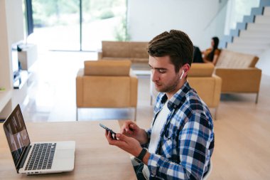 Modern bir oturma odasındaki masada oturan bir serbest çalışan, kulaklıklarıyla iş görüşmesi için bir laptop kullanan, arkadaşlarıyla sohbet eden ve eğlenen biri..