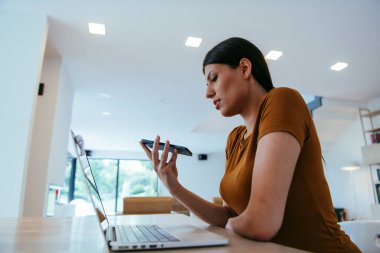 Oturma odasında dizüstü bilgisayar ve akıllı telefon kullanarak oturan bir kadın iş arkadaşları ve akrabalarıyla yaptığı görüntülü konuşmaya bakıyor. İş görüşmesi soruları yanıtlıyor