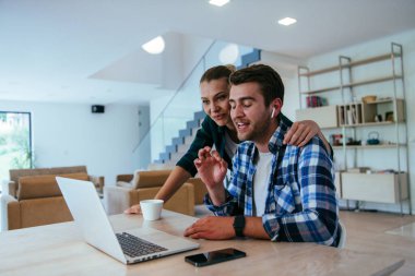 Genç evli bir çift, modern evlerinin oturma odasında otururken bir dizüstü bilgisayarla ebeveynleri, aileleri ve arkadaşlarıyla konuşuyor.