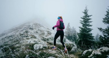 Soğuk ve sisli havada kayalık bir dağ yolunda koşan bir kadın sporcu, sıcak bir ceket, eldiven ve saç bandı giyiyor, dayanıklılık ve kararlılık sergiliyor.. 