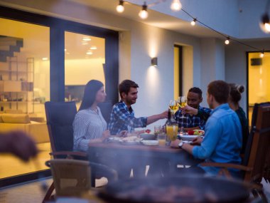Bir grup genç insan akşamları modern bir evin terasında yemek yiyorlar. Dostlar ve aile için eğlence. Tatil kutlamaları, barbekülü düğünler..