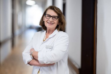 Tecrübeli ve kendinden emin yaşlı bir kadın doktor, gözlük takıyor ve hastane koridorunda duruyor, bilgeliği ve hasta bakımına bağlılığı temsil ediyor.