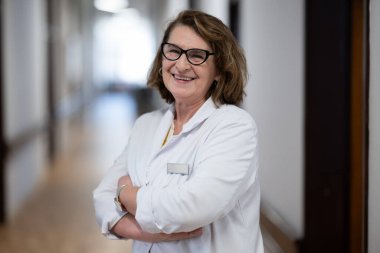 Tecrübeli ve kendinden emin yaşlı bir kadın doktor, gözlük takıyor ve hastane koridorunda duruyor, bilgeliği ve hasta bakımına bağlılığı temsil ediyor.