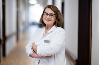 Tecrübeli ve kendinden emin yaşlı bir kadın doktor, gözlük takıyor ve hastane koridorunda duruyor, bilgeliği ve hasta bakımına bağlılığı temsil ediyor.