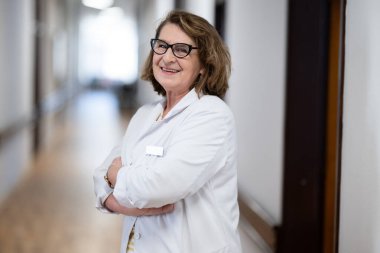 Tecrübeli ve kendinden emin yaşlı bir kadın doktor, gözlük takıyor ve hastane koridorunda duruyor, bilgeliği ve hasta bakımına bağlılığı temsil ediyor.