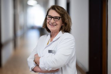 Tecrübeli ve kendinden emin yaşlı bir kadın doktor, gözlük takıyor ve hastane koridorunda duruyor, bilgeliği ve hasta bakımına bağlılığı temsil ediyor.
