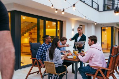 Bir grup genç insan akşamları modern bir evin terasında yemek yiyorlar. Dostlar ve aile için eğlence. Tatil kutlamaları, barbekülü düğünler..