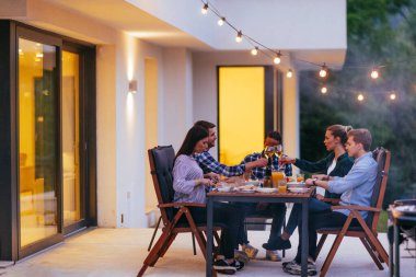 Bir grup genç insan akşamları modern bir evin terasında yemek yiyorlar. Dostlar ve aile için eğlence. Tatil kutlamaları, barbekülü düğünler..