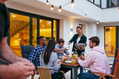 Bir grup genç insan akşamları modern bir evin terasında yemek yiyorlar. Dostlar ve aile için eğlence. Tatil kutlamaları, barbekülü düğünler..
