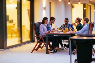 Bir grup genç insan akşamları modern bir evin terasında yemek yiyorlar. Dostlar ve aile için eğlence. Tatil kutlamaları, barbekülü düğünler..