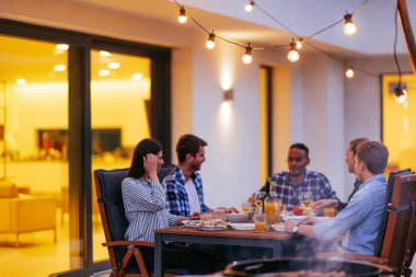 Bir grup genç insan akşamları modern bir evin terasında yemek yiyorlar. Dostlar ve aile için eğlence. Tatil kutlamaları, barbekülü düğünler..