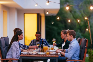 Bir grup genç insan akşamları modern bir evin terasında yemek yiyorlar. Dostlar ve aile için eğlence. Tatil kutlamaları, barbekülü düğünler..