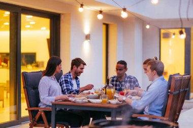 Bir grup genç insan akşamları modern bir evin terasında yemek yiyorlar. Dostlar ve aile için eğlence. Tatil kutlamaları, barbekülü düğünler..