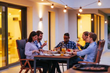 Bir grup genç insan akşamları modern bir evin terasında yemek yiyorlar. Dostlar ve aile için eğlence. Tatil kutlamaları, barbekülü düğünler..